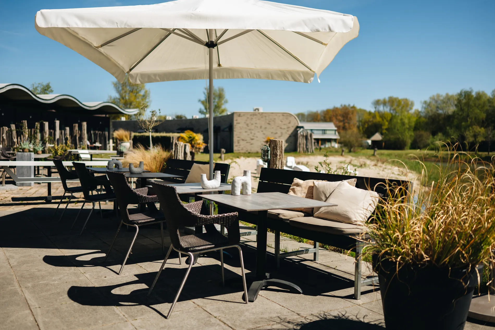 Moderne terrastafels en stoelen onder een grote witte parasol op het buitenterras, met op de achtergrond de natuur en het water.